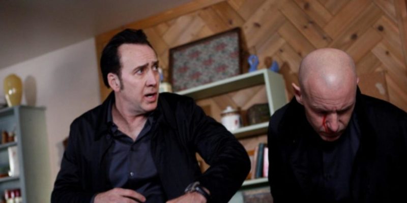 2017 Nicolas Cage Humanity Bureau - Nicolas Cage stands in a living room with a bald man.